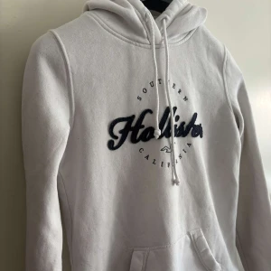 Vit hoodie från Hollister - Vit hoodie från Hollister med broderad logga i mörkblått på bröstet. Tröjan har huva med snörning, magficka och ribbade muddar. Perfekt för en chill och avslappnad stil. S/Xs
