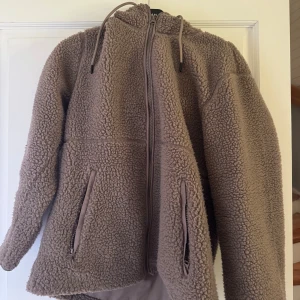 Beige teddyjacka med huva - Mysig beige teddyjacka med huva och dragkedja framtill. Jackan har två fickor med dragkedja och en avslappnad passform som är perfekt för kyliga dagar.