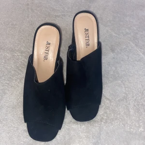 Svarta klackskor från JustFab - Svarta klackskor från JustFab med öppen tå och bred klack. Skorna har en stilren design med mockaliknande material och fyrkantig tå. Perfekta för dig som vill ha en trendig och bekväm sko med lite höjd.