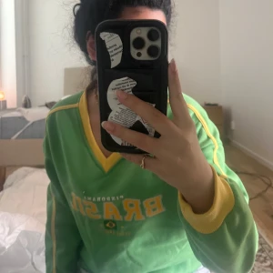Grön Brasil-tröja med gula detaljer - Säljer en grön sweatshirt med texten 'Brasil' och gula detaljer längs ärmar och krage. Tröjan har v-ringad hals och tryck framtill med brasiliansk flagga. Perfekt för dig som vill sticka ut med sportig vibe.