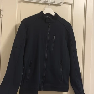 Mörkblå jacka från Koroshi Brand - Stilren mörkblå jacka från Koroshi Brand med hög krage och tre dragkedjeförsedda fickor framtill. Jackan har en clean look och är tillverkad i en mix av bomull, polyester och elastan för skön passform. Perfekt för dig som gillar minimalistisk stil.