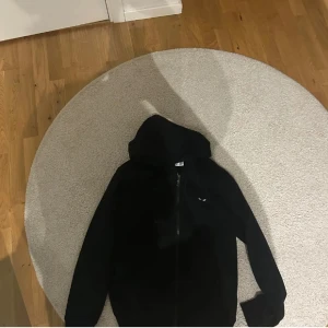 CDG Kofta - Svart kofta från Comme des Garçons Play med dragkedja och huva. På bröstet finns det ikoniska hjärtmotivet med ögon i svart. Långärmad och i mjukt material, perfekt för en chill och stilren look.