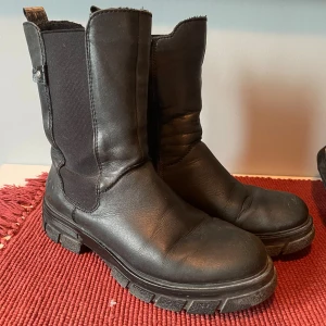 Svarta boots från Rieker i skinn - Tuffa svarta boots från Rieker i skinn med grov sula och dragkedja på insidan. Modellen har elastiska partier på sidorna för extra komfort och en robust look. Perfekta för höst och vinter när du vill ha både stil och funktion. Använd en säsong. Köparen betalar frakten. 