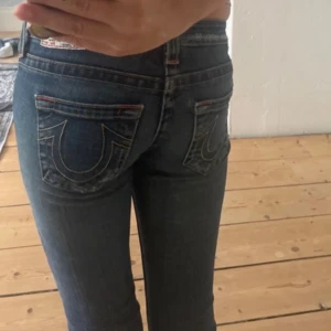 True Religion skinny jeans blå, stl 24 - Säljer ett par klassiska blå skinny jeans från True Religion med ikoniska bakfickor och kontrastsömmar. Jeansen har låg midja och är tillverkade i mjukt denim med snygg slitning och en liten reva på benet. Perfekt för dig som gillar streetstyle och vill sticka ut. 33 midjemått,80 innerbenslängd . Lågmidjade.