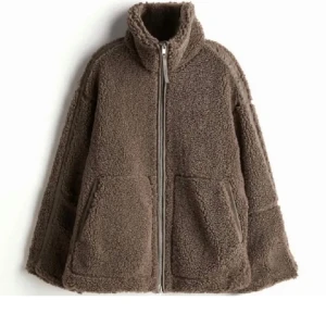 Brun teddyjacka i pile med dragkedja - Mysig brun teddyjacka med dragkedja framtill och hög krage. Perfekt för kyliga dagar med sin fluffiga och varma design. Jackan har två stora fickor framtill för extra komfort och stil.