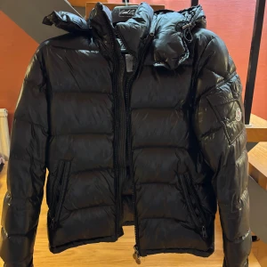 Svart Moncler Maya  - Svart pufferjacka från Moncler med glansig finish och huva. Jackan har Moncler-logga på ärmen, praktiska fickor med dragkedja och quiltad design. Perfekt för kalla dagar och har en modern, sportig look som sticker ut.