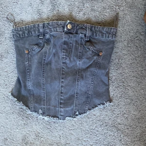 Grå jeanskorsett från Zara - Säljer en grå jeanskorsett från Zara i storlek S. Toppen har fransig nederkant, bälteshällor, fickdetaljer och dragkedja bak. Gjord i bomull med en cool denimlook och markerade sömmar. Perfekt att styla med höga byxor eller kjol.
