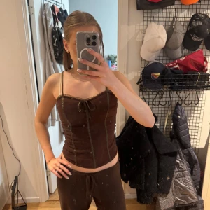 Brun korsett liknande linne  - Säljer en brun korsett liknande linne  med smala axelband och snygga snörningsdetaljer framtill. Toppen har markerade sömmar och en croppad passform som ger en cool siluett. Perfekt för dig som gillar trendiga och unika plagg med lite edge.