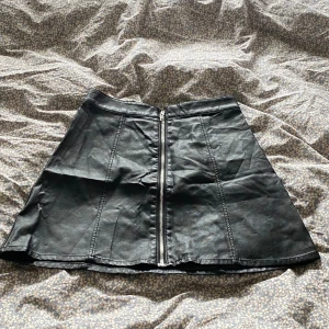 Svart kort kjol i skinnimitation - Snygg svart kort kjol i skinnimitation med markerade sömmar och dragkedja framtill. Kjolen har en lätt utsvängd form som ger en cool vibe och passar perfekt till en stickad tröja eller en tuff jacka. Perfekt för dig som gillar en edgy stil.