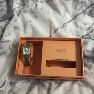 Guldig klocka från Regal med armband - Snygg guldig klocka från Regal med rektangulär urtavla och länkarmband. Urtavlan är vit med svarta romerska siffror och detaljer. Kommer i originalask tillsammans med matchande guldarmband. Perfekt accessoar för att lyfta din stil. Med helt set får man med köptes för 1000kr