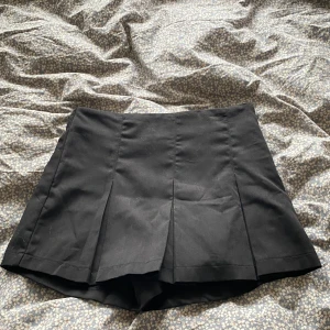 Svart plisserad kortkjol - Snygg svart kortkjol med plisserade detaljer framtill. Kjolen har en klassisk och stilren look som passar perfekt till sneakers eller boots. Enkel att matcha med olika toppar för en trendig outfit.