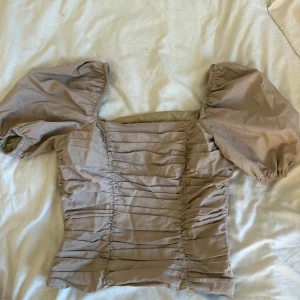 Beige puffärm blus från Chloé B x NA-KD - Supersöt beige blus från Chloé B x NA-KD med puffiga korta ärmar och snygga rynkade detaljer framtill. Kvadratisk halsringning och figurnära passform som ger en trendig siluett. Perfekt för dig som vill sticka ut med en romantisk vibe.