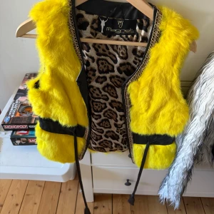 Gul pälsväst från Magnet Collection - Säljer en riktigt cool gul pälsväst med svart band och kedjedetaljer vid kragen. Insidan har leopardmönster som ger extra edge. Västen har snörning framtill och är perfekt för dig som vill sticka ut med färg och mönster.