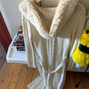 Vit lång cardigan jacka från DEX - Lång, stickad cardigan jacka i vit färg från DEX med mysig fuskpäls runt huvan. Snygga flätstickade detaljer, knappar framtill och praktiska fickor. Perfekt för lager-på-lager och chill dagar. Bälte i midjan för extra stilpoäng.