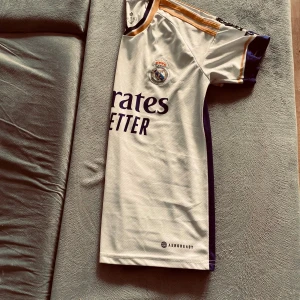 Real Madrid vit matchtröja Adidas - Snygg Real Madrid fotbollströja från Adidas i vit med marinblå detaljer och guldiga ränder på axlarna. Kortärmad modell med klubbmärke på bröstet och 'Emirates Fly Better' tryck. Tillverkad i lätt och ventilerande material med Aeroready-teknologi.