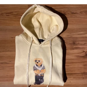 Gul hoodie Polo Ralph Lauren S - Snygg ljusgul hoodie från Polo Ralph Lauren med ikoniskt björntryck på bröstet. Tröjan har huva med snörning och ribbade muddar. Perfekt för dig som gillar streetstyle och vill ha något bekvämt men ändå stilrent.