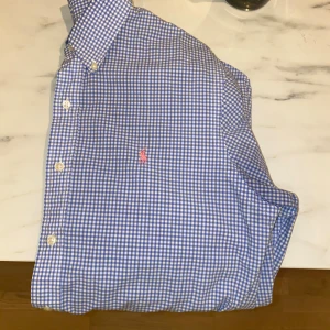 Blårutig skjorta från Polo Ralph Lauren - Klassisk blårutig skjorta från Polo Ralph Lauren med vit knappstängning och rosa broderad logga på bröstet. Skjortan har lång ärm, button-down krage och är tillverkad i mjuk bomull. Perfekt för en stilren och avslappnad look.