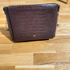 Brun axelväska i croco-mönstrat skinn - Säljer en snygg brun axelväska från  Anna Field. Den är mönstrad framsida och guldfärgade kedjedetaljer. Väskan har en stilren, fyrkantig form och justerbar axelrem i skinn. 