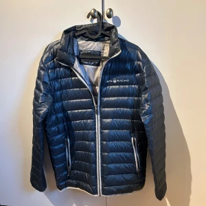 Mörkblå dunjacka från Sail Racing - Snygg mörkblå dunjacka från Sail Racing med quiltad design och vita dragkedjor. Jackan har hög krage, två fickor med dragkedja och logga på bröstet. Perfekt för kyliga dagar och riktigt lätt att bära.