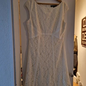 Vit spetsklänning från American Living - Superfin vit spetsklänning från American Living med klassisk rund halsringning och figurnära passform upptill. Klänningen har korta ärmar och vackert spetsmönster över hela plagget. Perfekt för sommarens alla tillfällen!