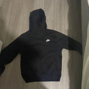 Svart Nike hoodie med huva - Svart hoodie från Nike med klassisk vit logga på bröstet. Tröjan har huva, lång ärm och en stor magficka framtill. Perfekt för dig som gillar en enkel och stilren look. Materialet är mjukt och skönt, troligtvis bomull eller bomullsblandning.Den har tappat lite färg och börjar bli röd annars är den helt perfekt.