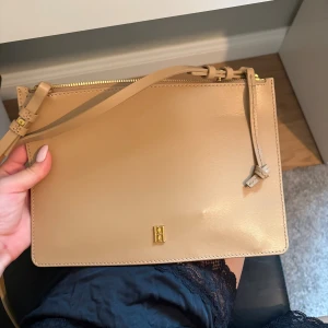 Beige axelväska från By Malene Birger - Stilren beige axelväska i slätt skinn från By Malene Birger. Väskan har guldfärgade detaljer, justerbar axelrem och dragkedja upptill. Minimalistisk design med en liten guldfärgad logga framtill. Perfekt för dig som gillar klassiska och enkla accessoarer.