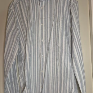 Randig skjorta från Riley Slim Fit XL - Snygg randig skjorta från Riley i slim fit-modell. Skjortan är vit med ljusblå ränder, har långa ärmar och knäppning framtill. Perfekt för dig som gillar en stilren och fräsch look. Tillverkad i bomull för en skön känsla.