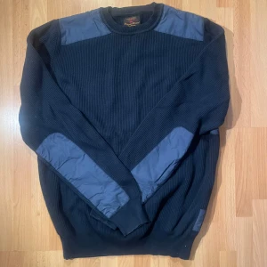 Mörkblå stickad tröja Barbour L - Säljer en mörkblå stickad tröja från Barbour i storlek L. Tröjan har ribbad struktur, förstärkta partier på axlar och armbågar i tyg samt rund hals. Perfekt för dig som gillar klassisk och stilren look. Pyttelitet hål i ryggen tog en bra stund innan jag hittade det. Märks inte alls! Över 20 år gammal