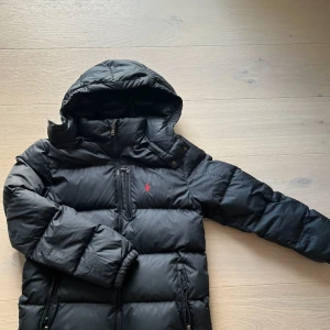 Svart dunjacka från Polo Ralph Lauren - Svart dunjacka från Polo Ralph Lauren med huva, dragkedjor och röd broderad logga på bröstet. Jackan är vadderad och har flera fickor med dragkedja. Perfekt för kalla dagar och riktigt snygg streetstil. Finns några tecken på användning på insidan av jackan men inget som syns på utsidan.