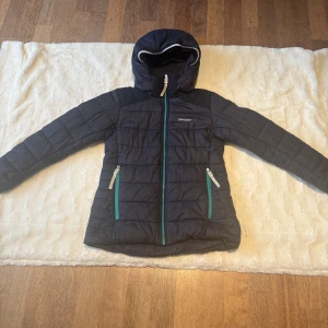 Blå quiltad pufferjacka Didriksons 140 - Blå quiltad pufferjacka från Didriksons1913 med huva och turkosa dragkedjor. Jackan är tillverkad i 100% polyester och har två fickor framtill. Perfekt för kyliga dagar och har en stilren look med diskret logga på bröstet. Storlek 140. Liten reva på baksidan (se bild 2). 