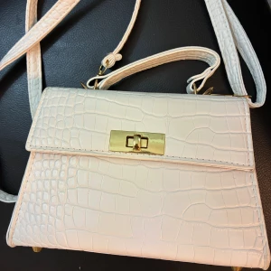 Vit croco handväska med gulddetaljer - Snygg vit handväska i croco-mönstrat skinnimitation med guldfärgade metalldetaljer och lås. Väskan har både handtag och axelrem, samt röd insida för en extra touch. Perfekt för dig som vill ha en stilren och trendig accessoar.