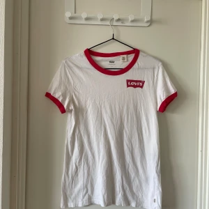 Vit Levi's t-shirt med röda detaljer - Vit t-shirt från Levi's med klassisk röd logga på bröstet och röda ribbade kanter vid hals och ärmslut. Modellen är kortärmad och har en avslappnad passform. Tillverkad i mjuk bomull, perfekt för en clean och sportig look.