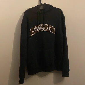 Svart hoodie Axel Arigato L - Svart hoodie från Axel Arigato i storlek L med vit broderad text 'ARIGATO' på bröstet. Klassisk modell med huva, snörning och känguruficka. Tillverkad i 100% bomull för en skön och avslappnad vibe.