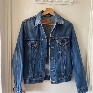 Klassisk jeansjacka från Levi's, M - Snygg blå jeansjacka från Levi's i klassisk modell med bruna knappar och två bröstfickor med lock. Jackan har raka linjer, krage och är tillverkad i slitstarkt denim. Perfekt för dig som vill ha en tidlös och cool look.