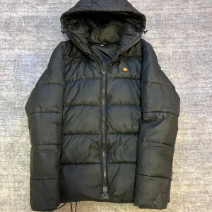 Svart pufferjacka från Ellesse - Säljer en svart pufferjacka från Ellesse i mycket bra skick. Jackan har en dragkedja framtill och en mysig huva. Perfekt för kalla dagar och håller dig varm och bekväm. Den har en klassisk pufferstil med quiltade paneler. Jag har bara haft jackan i mindre än ett år. Så den är fortfarande på topp skick. Kvitto finns också👍