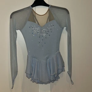 Isblå konståkningsklänning Jerry's - Superfin isblå konståkningsklänning från Jerry's med långa meshärmar, glittriga stenar och volangkjol. Klänningen har snygg v-ringning med meshdetaljer och är dekorerad med silver- och blå strass framtill. Materialet är stretchigt och följsamt för maximal rörelsefrihet.