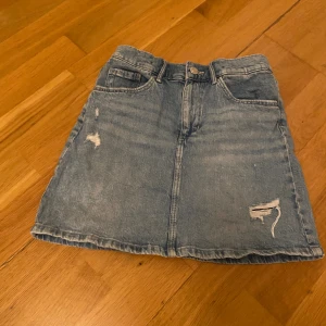 Ljusblå jeanskjol från &Denim - Snygg ljusblå jeanskjol från &Denim med slitna detaljer och klassisk femficksdesign. Kjolen har knapp och dragkedja framtill samt bälteshällor. Perfekt för dig som gillar en avslappnad och trendig stil.