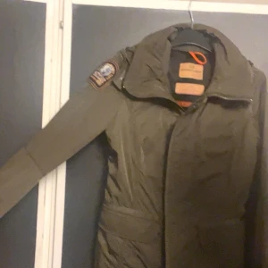Grön Parajumpers parkajacka med patch - Säljer en grön parkajacka från Parajumpers med snygg patch på ärmen och orange detalj vid halsen. Jackan har huva, dragkedja och knappar framtill samt bälte i midjan för schysst passform. Perfekt för kalla dagar och riktigt stilren look.