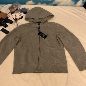 Grå zip hoodie från Polo Ralph Lauren - Grå hoodie från Polo Ralph Lauren med dragkedja framtill och klassisk huva. Tröjan har broderad logga på bröstet och är tillverkad i mjuk bomullsblandning. Perfekt för chill dagar och enkel att matcha med jeans eller joggers. Kan gå ner i pris.