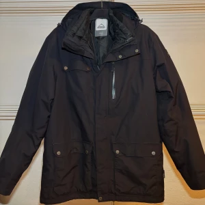 Svart parkajacka från McKinley L - Svart parkajacka från McKinley i storlek L. Jackan har huva med dragsko, flera fickor med tryckknappar och en bröstficka med dragkedja. Perfekt för kyliga dagar med sin långa passform och praktiska detaljer.