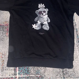Svart sweatshirt med nalletryck - Svart sweatshirt från Chaseone med ett stort tryck av en grå nallebjörn och en vit krona på bröstet. Tröjan har rund hals, ribbade muddar och är långärmad. Perfekt för dig som gillar streetstyle och coola prints.