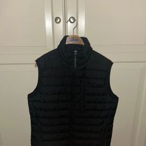 Svart dunväst från Peak Performance - Snygg svart dunväst från Peak Performance med hög krage och dragkedja framtill. Västen har en quiltad design och en bröstficka med dragkedja. Perfekt att slänga över en hoodie eller tröja när det är kyligt ute.