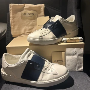 Valentino Garavani Open sneakers vit/blå - Säljer ett par Valentino Garavani Open sneakers i vitt skinn med bred marinblå läderdetalj över mitten. Klassisk rund tå, vita snören och ikoniska nitar baktill. Kommer med originalkartong, dustbag och extra skosnören. Perfekta för dig som gillar lyxiga och stilrena sneakers. Skorna är köpta i början på sommaren på Valentino som ligger i illum i Köpenhamn, skorna är sällan använda då jag har varit väldigt rädd om de. Om det är något mer ni undrar så är det bara att skriva, mvh Otto 😃