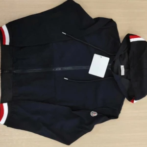 Svart hoodie från Moncler med ränder - Svart hoodie från Moncler med dragkedja, huva och snörning. Ärmar och huva har snygga vita och röda ränder. Moncler-logga på ena ärmen. Tillverkad i mjuk bomull, perfekt för en chill och stilren look. NY TRÖJA ALDRIG ANVÄND!