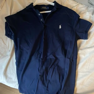 Marinblå pikétröja från Ralph Lauren - Säljer en marinblå pikétröja från Ralph Lauren i custom fit med klassisk krage och knappar. Tröjan har det ikoniska broderade polospelar-loggan på bröstet och är gjord i mjuk pima stretch mesh för extra komfort. Perfekt för en clean och stilren look.