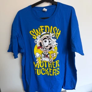 Turbojugend Sverige T-shirt xxl - Blå t-shirt med stor, färgglad och galen tecknad print på framsidan med texten 'Swedish Mother Fuckers' i gult. Motivet visar en skruvad karaktär med öl och skallar, perfekt för dig som gillar unika och roliga plagg. Rund hals och korta ärmar.