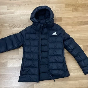 Svart pufferjacka från Adidas - Svart pufferjacka från Adidas med huva och vit logga på bröstet. Jackan är quiltad och har dragkedja framtill. Perfekt för kalla dagar och har en clean, sportig look. Tillverkad i syntetmaterial och har långa ärmar.