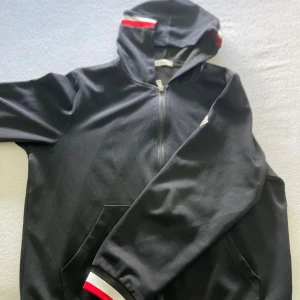 Svart hoodie med röd och vit detalj - Svart hoodie med dragkedja och huva. Snygga röda och vita ränder på huvan och vid ärmsluten ger en sportig vibe. Praktiska fickor framtill och mjukt material, perfekt för chill dagar eller när du vill ha en enkel streetstyle.