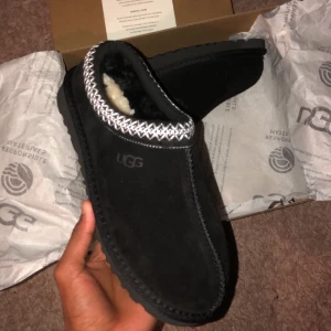 Svarta UGG i mocka - Svarta slip-in skor från UGG med mjuk insida i fårull och ovandel i mocka. Skorna har dekorativt mönster i vitt runt öppningen och en robust, mönstrad sula för extra grepp. Perfekta för att hålla fötterna varma och bekväma under kyliga dagar.