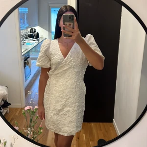 Vit omlottklänning från Kappahl - Supersöt vit omlottklänning från Kappahl med puffärmar och strukturmönster i tyget. Klänningen har knyt i sidan och v-ringning, vilket ger en riktigt fin siluett. Perfekt för sommarens alla tillfällen!